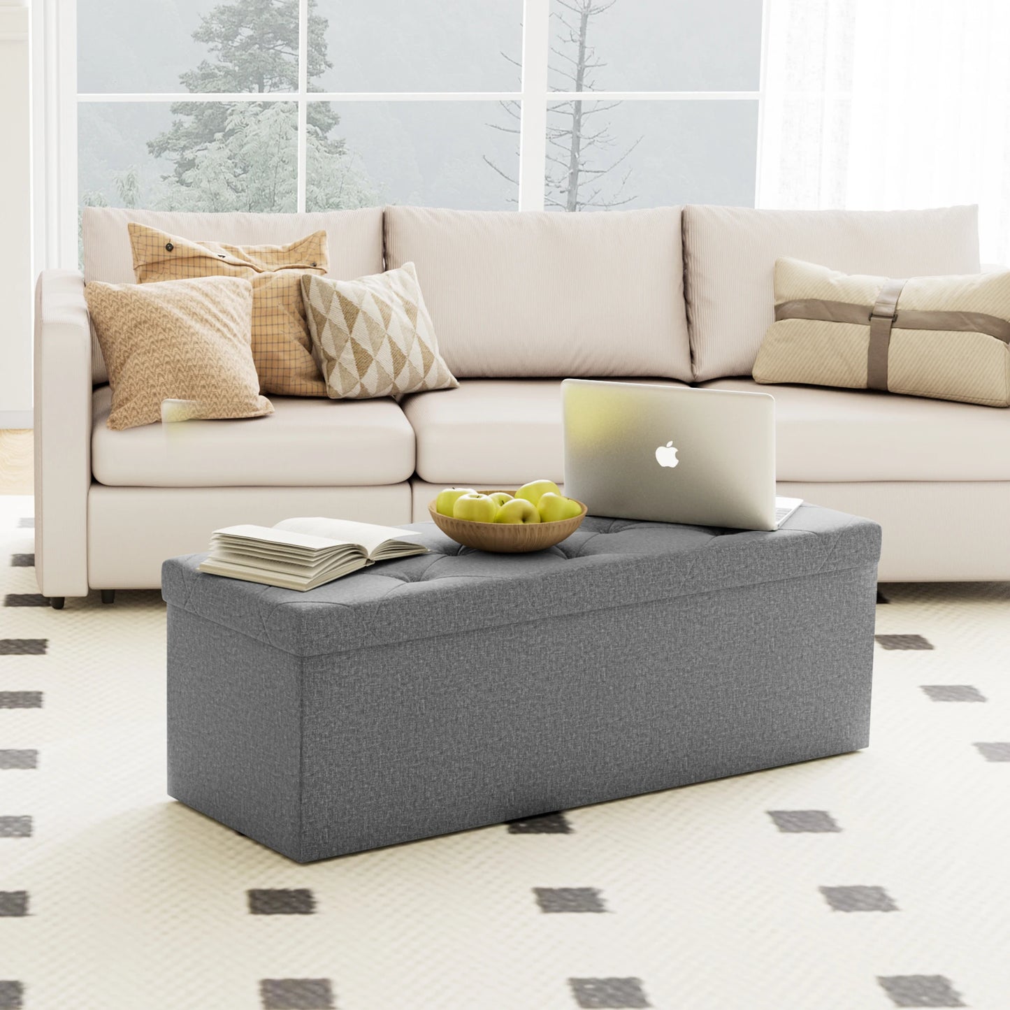 Gray ottoman with a laptop, book, and bowl of apples on top, in front of a white sofa with decorative pillows.
